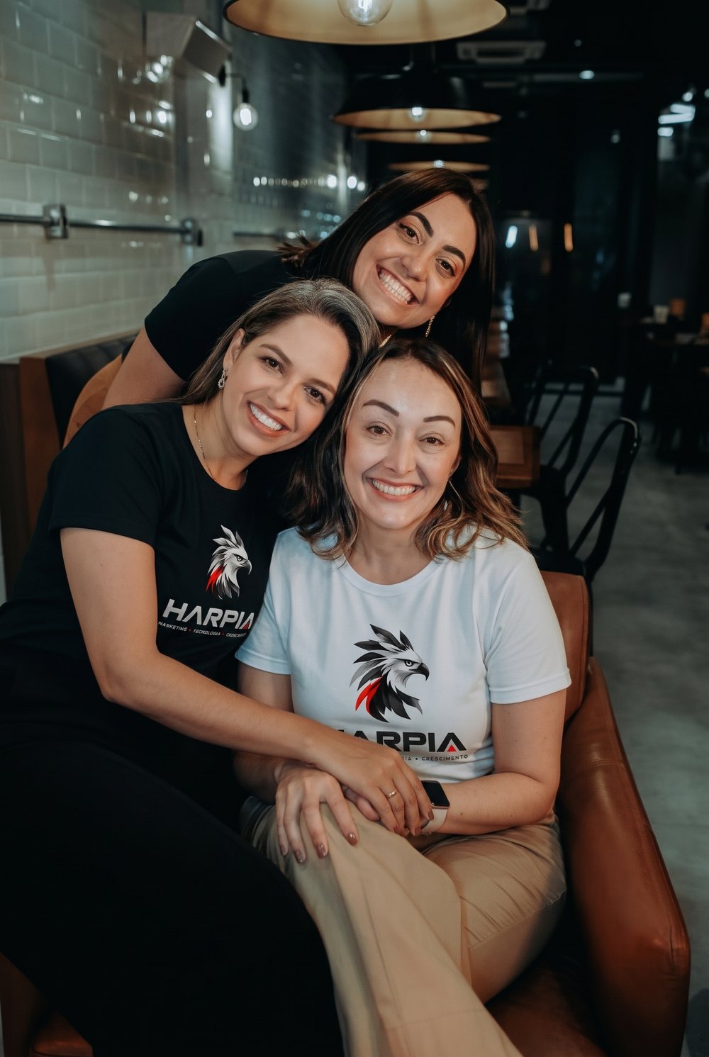 Três integrantes do time Harpia abraçadas e sorrindo, com camisetas da marca